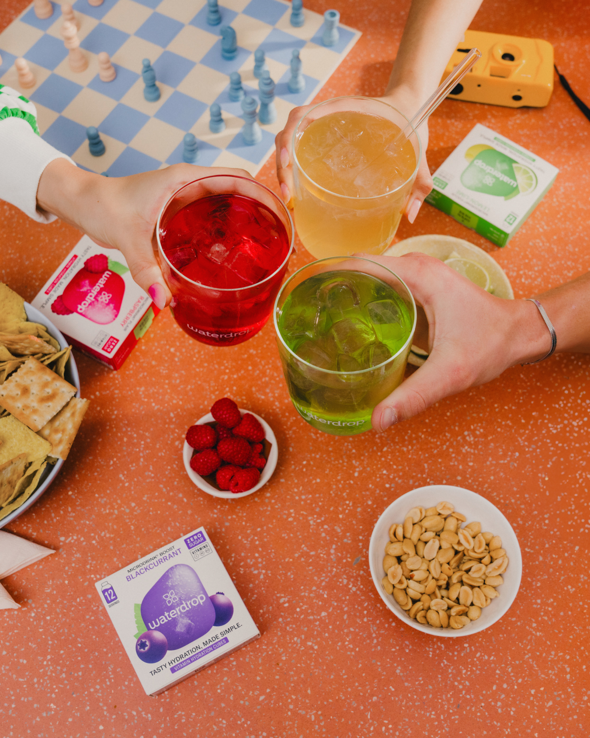 Hands holding glasses of drinks around a table with snacks, featuring a bowl of raspberries, highlighting the refreshing Raspberry-flavored waterdrop® Microdrink.